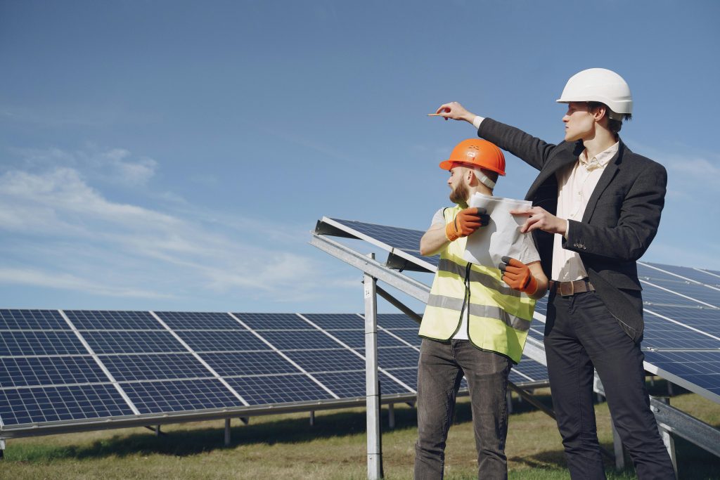 Young businessman in formal outfit and foreman in yellow vest looking away while discussing working process against solar panels